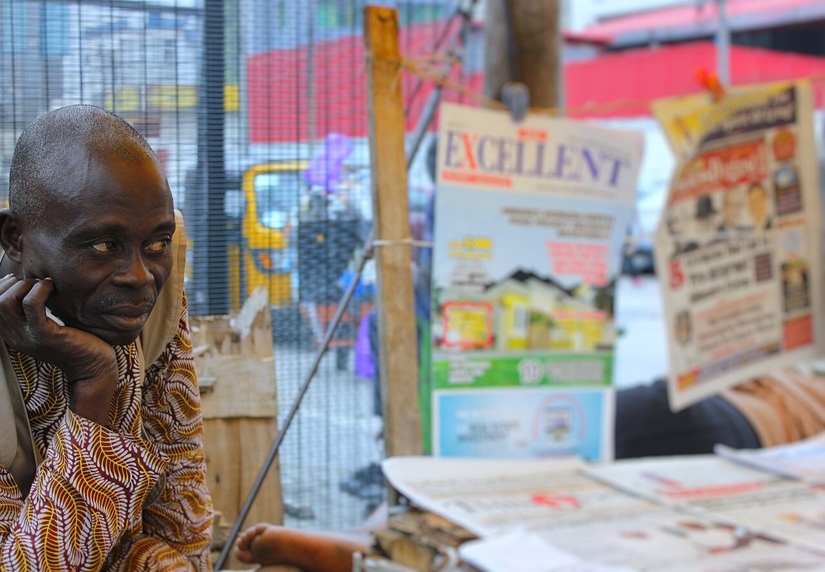 A newspaper vendor at his street stand in Nigeria, with the day’s papers laid out and pegged up beside him